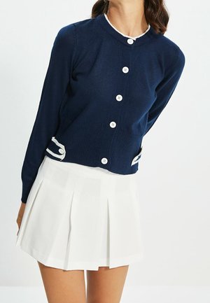 Femme portant un cardigan bleu marine boutonné avec un bord blanc et une jupe plissée blanche sur un fond uni.