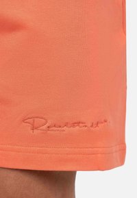 Oranje sportshorts gemaakt van rekbare stof. Heeft een geborduurd logo in een contrasterende kleur nabij de zoom. Soepele textuur, aansluitend ontwerp.