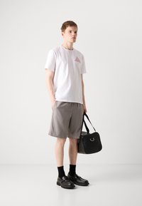 White cotton t-shirt with red triangular logo, gray shorts, black leather shoes with chunky soles, carrying black bag with silver hardware.