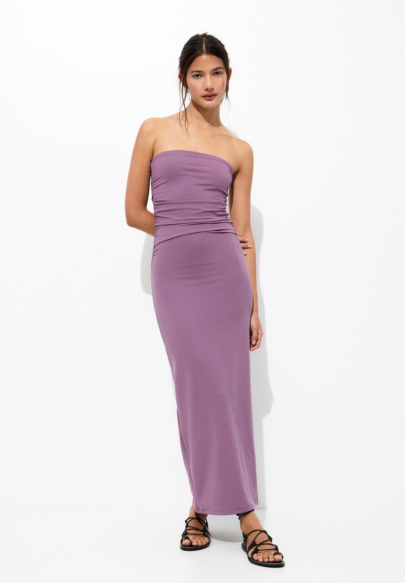 Robe maxi sans bretelles violette avec une silhouette ajustée, des détails froncés à la taille et une texture lisse. Assortie à des sandales noires à lanières.