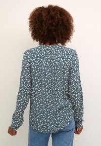 Chemise à manches longues en bleu-vert avec des motifs abstraits blancs et roses, dotée d'un col rond et d'un ourlet incurvé, fabriquée en tissu doux.