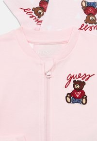 Ροζ μπουφάν με φερμουάρ και κεντητό σχέδιο αρκούδας με κόκκινο πουκάμισο. Μαλακό ύφασμα, στρογγυλή λαιμόκοψη, ορατή ετικέτα μάρκας.