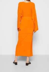 Orange långärmad klänning med figursydd midja och utsvängda ärmar, tillverkad av mjukt tyg som flödar ner till en rak nederkant med subtil textur.