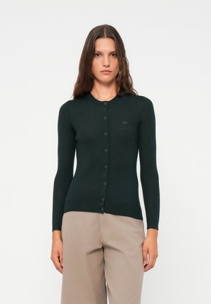 Mørkegrønn ribbestrikket cardigan med rund hals, lange ermer og knappelukking foran med matchende knapper. Utsmykket med en liten logo.