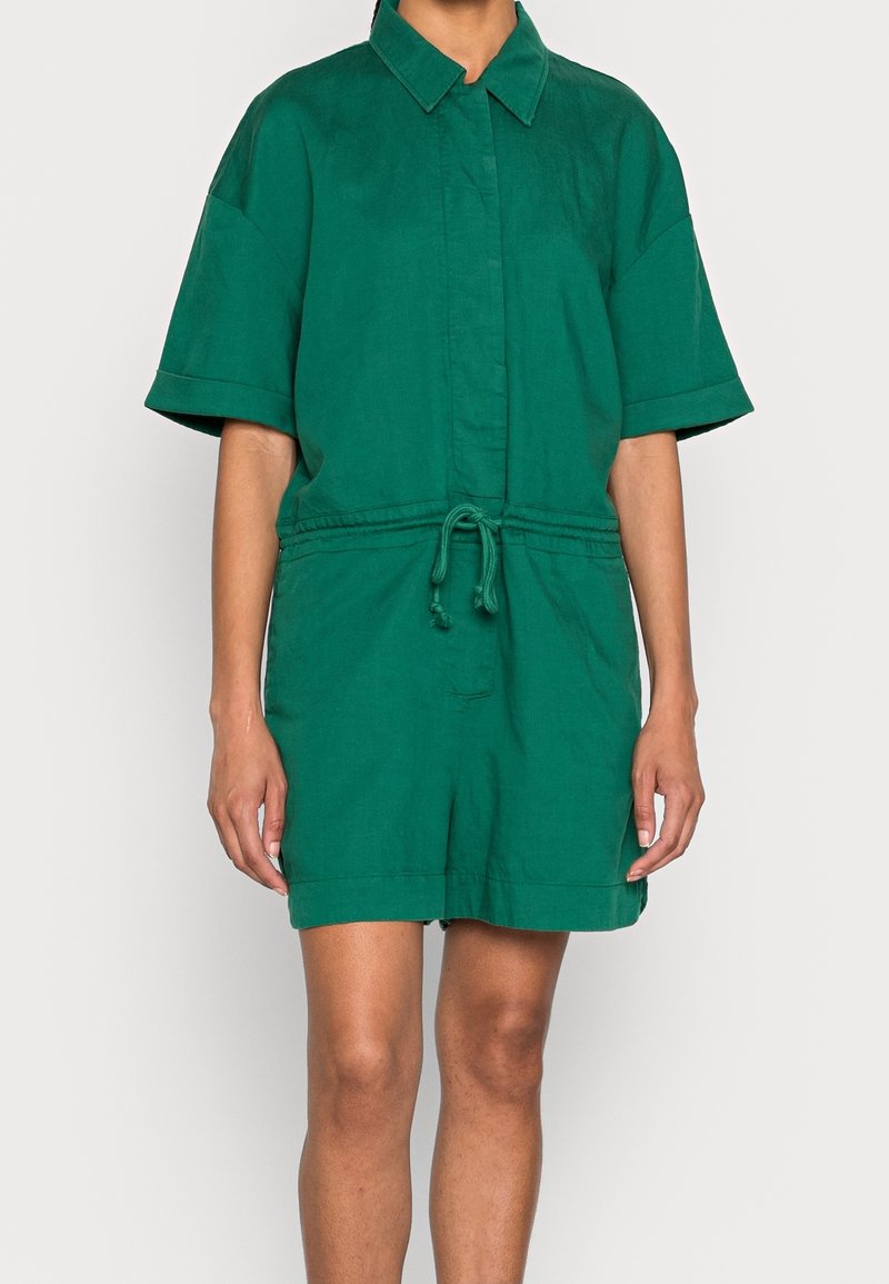 Personne portant une combinaison verte à manches courtes avec col, taille ajustable à cordon devant, et short jusqu’aux genoux sur un fond uni.
