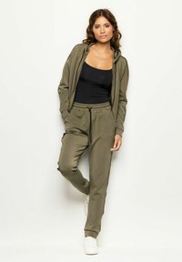 Oliwkowy dres wykonany z gładkiego materiału; zawiera rozpinaną bluzę na suwak i spodnie z elastycznym pasem z czarnymi akcentami po bokach. Całość dopełniają białe sneakersy.