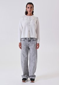 Cardigan bianco a maglia con ricamo floreale sulle spalle, cinque bottoni, abbinato a jeans larghi grigio chiaro e sneakers nere.