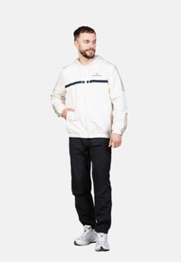 Chaqueta con capucha de color beige claro con una franja azul marino, bolsillos frontales y puños elásticos, combinada con pantalones negros y zapatillas blancas.
