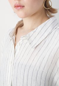 Frau trägt ein weißes, leichtes, gestreiftes Hemd mit Knöpfen und goldenen Creolen, gezeigt von Hals bis zur Mitte der Brust, neutraler Hintergrund.
