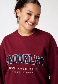 Bordeaux sweatshirt met ronde hals met "BROOKLYN" in marineblauwe letters en "NEW YORK CITY ATHLETIC DEPT." eronder in het wit. Zachte textuur.