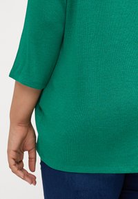 Grønn strikket topp med albue-lange ermer, teksturert stoff, avrundet kant og romslig passform. Hånd hviler på siden, iført mørk denim jeans.