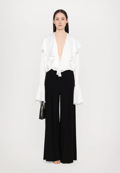 Witte blouse met ruches aan de kraag, diepe V-hals en lange mouwen; gecombineerd met losse zwarte broek; model met een zwarte gestructureerde handtas.