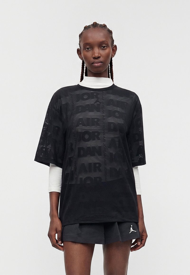 Jeune personne aux cheveux tressés portant un t-shirt noir oversize Jordan Air par-dessus un t-shirt blanc à manches longues et un short noir avec un logo blanc.