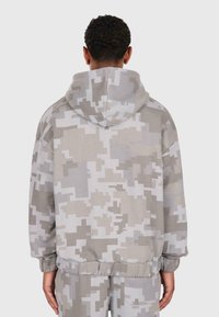 Bărbat îmbrăcat într-un hanorac cu camuflaj pixelat gri și alb, împreună cu pantaloni asortați, văzut din spate pe un fundal simplu.