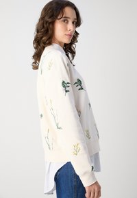 Kremfarget hettegenser med blomsterbroderi i grønt og gult. Den har en avslappet passform og et lagdelt utseende med en hvit skjorte under.