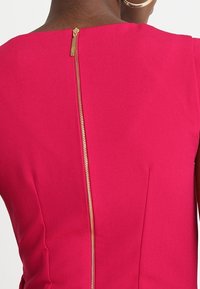 Robe ajustée en tissu rose vif, avec une texture lisse et une fermeture éclair dorée au centre du dos, agrémentée de détails de coutures discrètes.