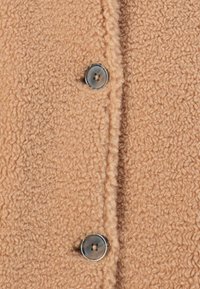 Beiger, flauschiger Stoff mit strukturiertem Finish, ausgestattet mit einem silbernen Knopfverschluss. Der Stoff hat ein weiches, warmes Aussehen mit sichtbaren Nähten.