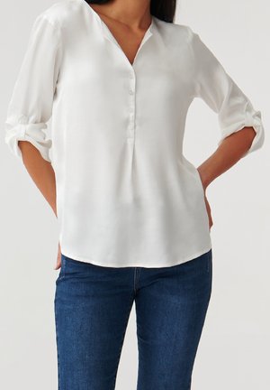 Hvid bluse i silkeagtigt materiale med V-udskæring, knaplukning foran og trekvartlange opsmøgede ærmer. Stylet med mørkeblå jeans.