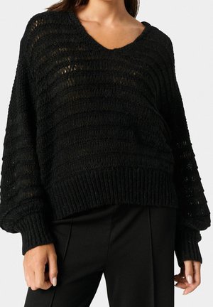 Czarny sweter z dzianiny ma dekolt w serek, luźny krój oraz strukturalny, prążkowany wzór z szerokimi rękawami. Nosi się go na czarnych spodniach.