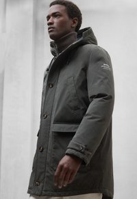 Oliwkowo-zielona parka z kapturem, dwoma przednimi kieszeniami i brązowymi guzikami; teksturowana tkanina z ekologiczną metką na rękawie.