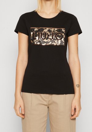 Kobieta w czarnej koszulce z błyszczącym brązowym logo "LIU·JO" i beżowych spodniach, stojąca na jednolitym tle.