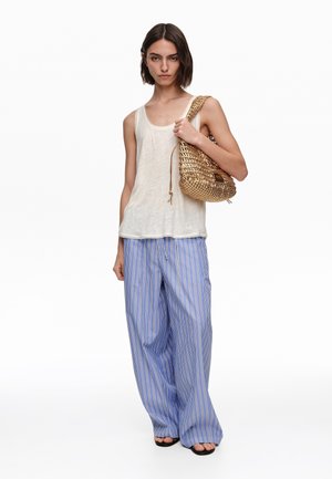 Kobieta nosząca szerokie niebiesko-białe spodnie w paski i białą bluzkę bez rękawów, trzymając na ramieniu plecioną złotą torebkę.