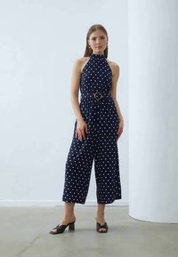 Morski jumpsuit z belimi pikami, zasnova z ovratnikom za halter, elastičen pas z okroglo pasom, široki skrajšani hlačnici, obut s črnimi mule sandalčki.