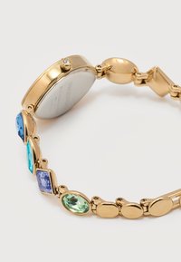 Χρυσό ρολόι χειρός με πολύχρωμους πολύτιμους λίθους στο λουρί, με στρογγυλό καντράν και ένα μικρό πολύτιμο λίθο στην κορώνα.