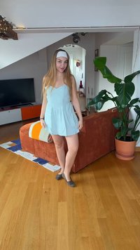 Jeune femme en robe bleu clair et bandeau debout sur un sol en bois près d'un canapé orange et d'une grande plante en pot dans un salon.