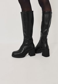 Bottes noires en cuir montantes avec des talons carrés, présentant une texture lisse, des panneaux élastiques sur les côtés et une fermeture éclair sur le côté pour un enfilage facile.
