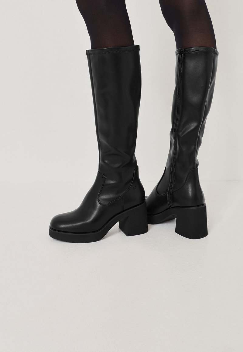 Bottes noires en cuir montantes avec des talons carrés, présentant une texture lisse, des panneaux élastiques sur les côtés et une fermeture éclair sur le côté pour un enfilage facile.