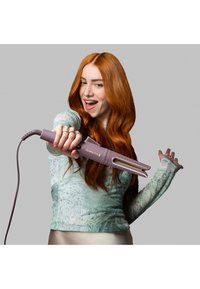 Varmeverktøy i lys lilla med gullfarger, med en buet form og teksturert grep, holdt av en person med langt rødt hår.