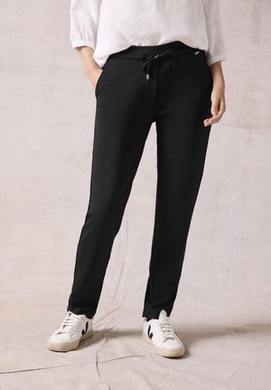 Person trägt schwarze Hose mit Kordelzug, weiße Sneakers und ein weißes Langarmshirt, steht vor neutralem Hintergrund.