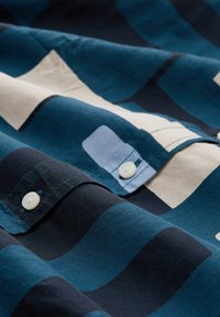 Camicia di cotone blu scuro e crema con motivi a onda. Presenta bottoni e pannelli in tessuto a contrasto. Tessuto liscio.