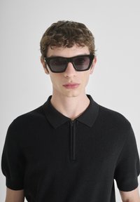 Crna polo majica kratkih rukava izrađena od rebrastog materijala, s ovratnikom na zatvarač. Model nosi uglaste crne sunčane naočale.