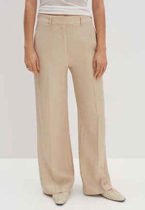 Pantalones beige de pierna ancha hechos de una tela suave, con un pliegue frontal y un cierre oculto. Combinados con zapatos planos de color crema.
