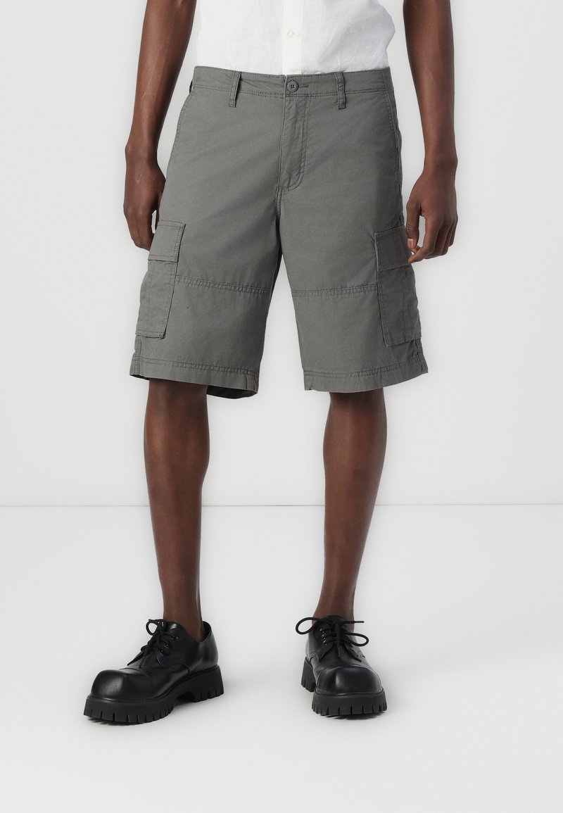 Graue Cargo-Shorts mit mehreren Taschen, aus strapazierfähigem Stoff, mit geradem Schnitt und genähten Details. Getragen mit schwarzen Schuhen.