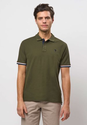 Mand iført olivengrøn polo med navy og hvide striber på ærmerne, stående foran en lysegrå baggrund.