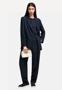 Traje azul marino a medida con pantalones sueltos y una americana, combinado con una blusa azul marino y un bolso blanco. Tela suave y diseño estructurado.
