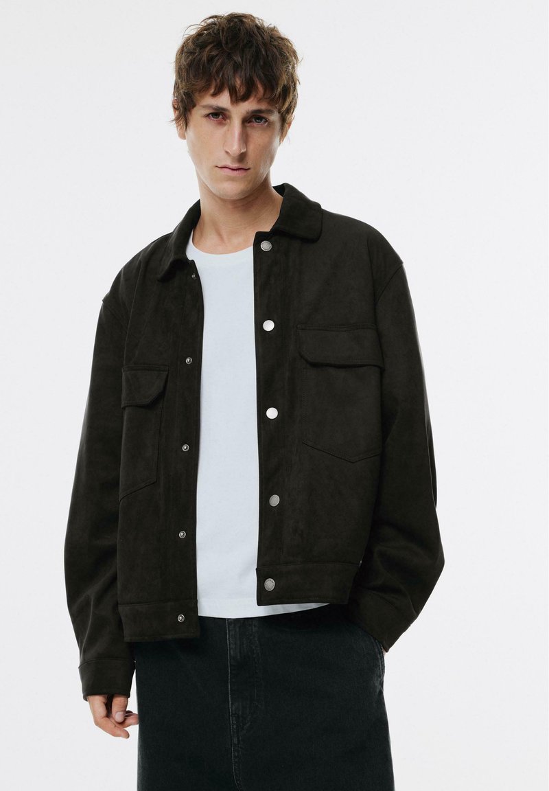Zwarte suède jas met drukknoopjes, twee voorzakken en een kraag, gelaagd over een wit shirt en gecombineerd met donkere broek.