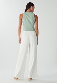 Top verde senza maniche abbinato a pantaloni bianchi a gamba larga. Il top presenta una texture liscia, mentre i pantaloni hanno una caduta fluida con un design minimalista.