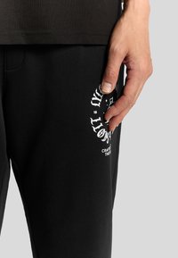 Zwarte sweatpants van zachte stof, met een witte grafische print en tekst op het been. Hand op zak voor detail.