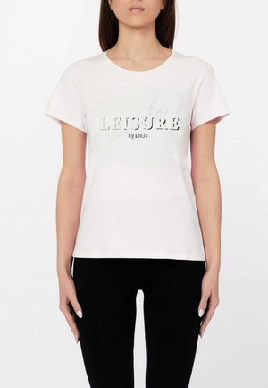 Donna che indossa una T-shirt bianca a maniche corte con la scritta "LEISURE by Liu Jo" sul davanti, abbinata a pantaloni neri, in piedi davanti a uno sfondo bianco.