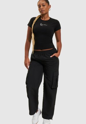 T-shirt nera aderente con logo bianco, abbinata a pantaloni cargo neri larghi con tasche laterali. Il modello indossa sneakers bianche.