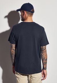 Camiseta de algodón azul marino de manga corta, con escote redondo y un ajuste relajado, combinada con pantalones beige, mostrando intrincados tatuajes en los brazos.