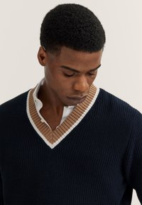 V-Ausschnitt Pullover in dunkelblau mit gerippter Textur, mit einem kontrastierenden Kragen in Beige und Weiß. Der Stoff wirkt weich und gestrickt.