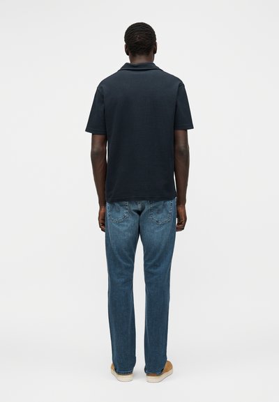 Homme de dos, portant un polo noir à manches courtes, un jean bleu et des baskets marron clair sur un fond uni.