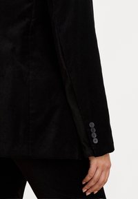 Blazer in velluto nero caratterizzato da una texture liscia, una forma sartoriale e una fila di sei bottoni neri sul polsino per la chiusura.