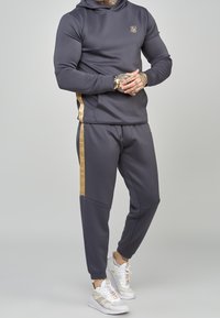 Felpa sportiva grigio antracite con logo dorato, abbinata a pantaloni jogger coordinati con striscia laterale dorata e dettagli testurizzati.