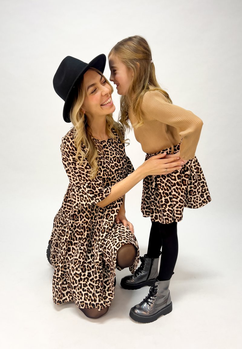 Leopardenmuster-Kleider, eines knielang und eines ausgestellt, kombiniert mit schwarzen Leggings und metallic-grauen Stiefeln, vor einem weißen Hintergrund.